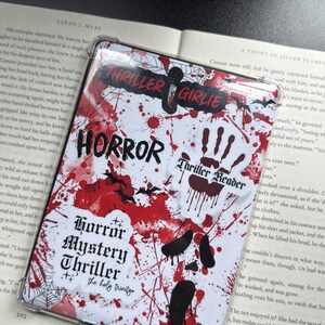 May include: A clear phone case with a white background, splattered with red paint and black accents. The case features the words "HORROR," "Thriller Girlie," and "Horror Mystery Thriller" in a spooky font. A handprint and bat silhouettes are also visible.