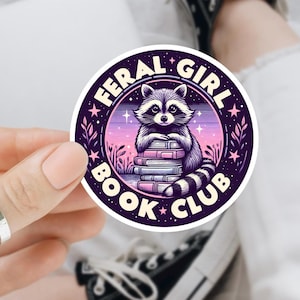 May include: A round sticker featuring a raccoon sitting on a stack of books. The sticker has a purple and pink color scheme with the words "FERAL GIRL BOOK CLUB" in a circular design. The raccoon has a black and white striped tail.