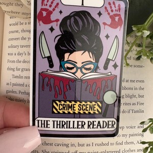 May include: A rectangular sticker featuring a tarot card design with a person in blue glasses reading a book covered in red blood drips. The sticker includes two knives, two red handprints, a magnifying glass, and the text CRIME SCENE and THE THRILLER READER.