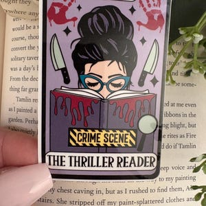 May include: A rectangular sticker designed like a tarot card featuring a person with a dark bun and blue glasses reading a book. The book has red blood-like drips, a yellow and black CRIME SCENE label, and the text THE THRILLER READER at the bottom.