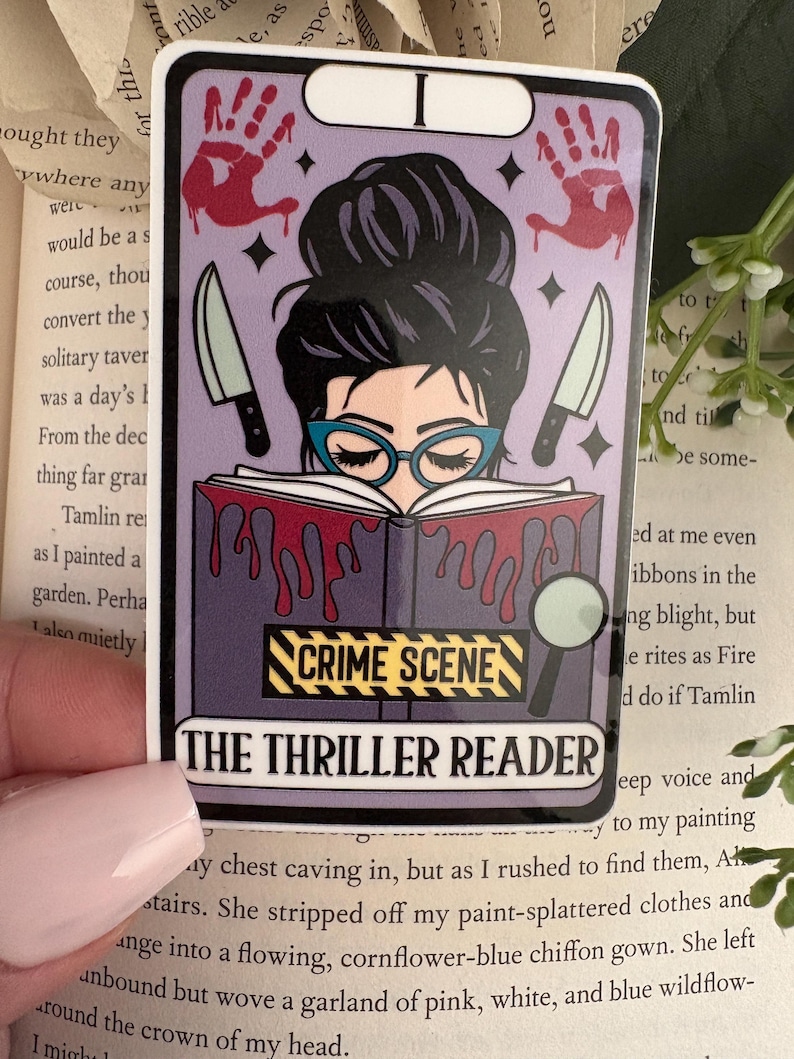 May include: A rectangular sticker designed like a tarot card featuring a person with a dark bun and blue glasses reading a book. The book has red blood-like drips, a yellow and black CRIME SCENE label, and the text THE THRILLER READER at the bottom.