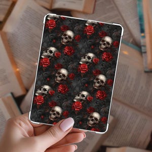Puede incluir: Una funda para tableta con un patrón repetido de calaveras plateadas y rosas rojas sobre un fondo gris oscuro. La funda tiene un borde plateado y se sostiene en una mano.