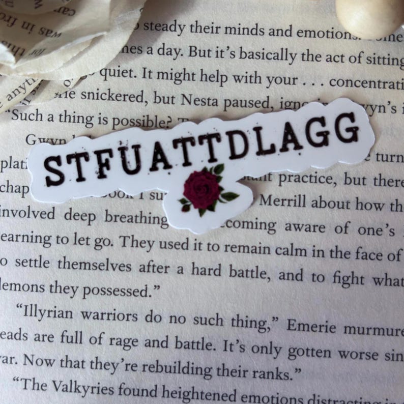 May include: A white die-cut sticker featuring the black text STFUATTDLAGG in a bold serif font. Below the text, a small, detailed illustration of a dark red rose with green leaves is centered, adding a floral accent to the sticker design.