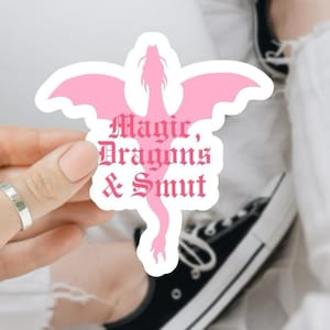 May include: A pink sticker with a dragon design and the words "Magic, Dragons & Smut" in a gothic font. The sticker is held by a person. The dragon has outstretched wings and a long, serpentine tail.