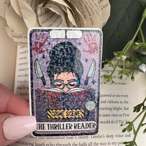 May include: A rectangular holographic sticker featuring a person with a dark bun hairstyle and teal glasses reading a book. The book has red drips, two knives, and the text CRIME SCENE and THE THRILLER READER. Red handprints are visible above.