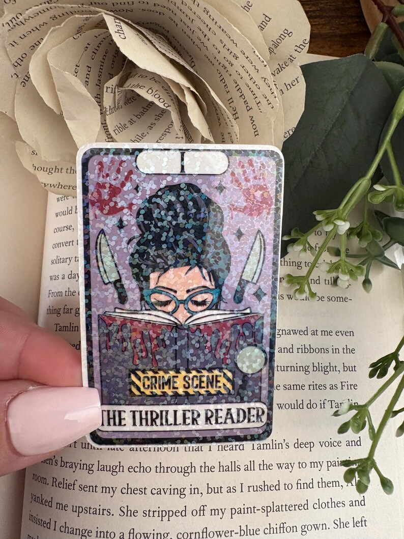 May include: A rectangular holographic sticker featuring a person with a dark bun hairstyle and teal glasses reading a book. The book has red drips, two knives, and the text CRIME SCENE and THE THRILLER READER. Red handprints are visible above.
