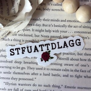 May include: A white die-cut sticker featuring the black text STFUATTDLAGG with a small, detailed dark red rose illustration positioned below the letters. The sticker has a scalloped edge and is placed against a background of printed book pages.