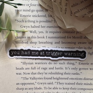 以下が含まれることがあります： 黒いステッカーに「you had me at trigger warning」と書かれた本のページのクローズアップ。ページのテキストは、イリュリアの戦士とその戦闘における高揚した感情について論じています。