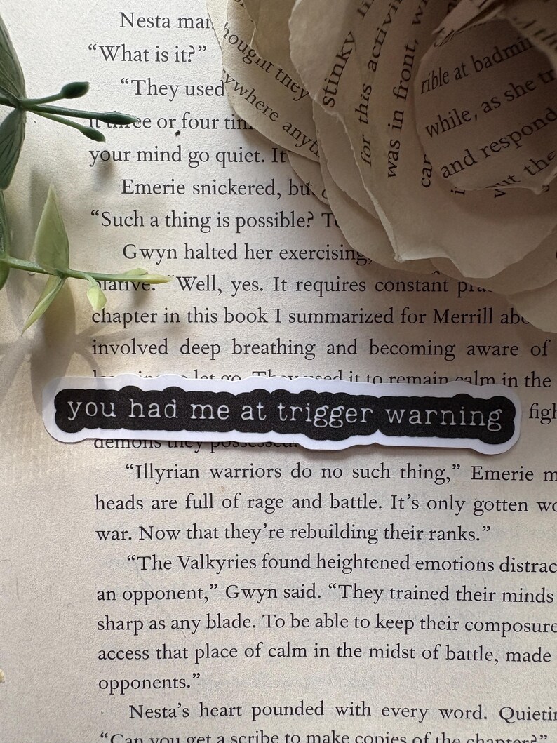 以下が含まれることがあります： 黒いステッカーに「you had me at trigger warning」と書かれた本のページのクローズアップ。ページのテキストは、イリュリアの戦士とその戦闘における高揚した感情について論じています。