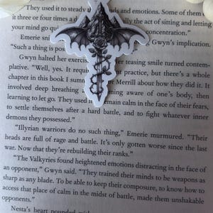 May include: A die-cut sticker featuring a central sword pointed downward, entwined with a thorny vine and a blooming rose. The sword hilt is topped with a mountain peak and flanked by large, detailed bat wings, all rendered in black and white ink.