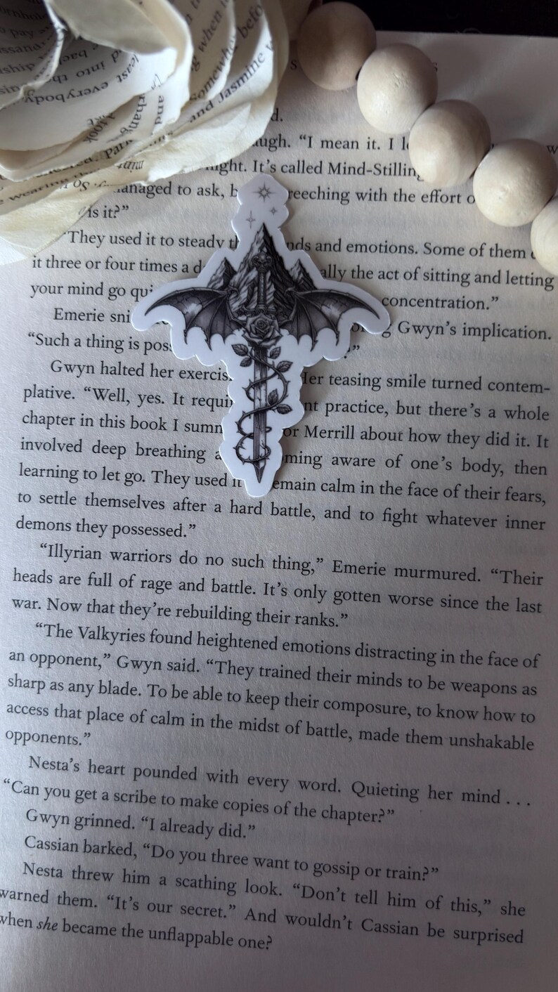 May include: A die-cut sticker featuring a central sword pointed downward, entwined with a thorny vine and a blooming rose. The sword hilt is topped with a mountain peak and flanked by large, detailed bat wings, all rendered in black and white ink.