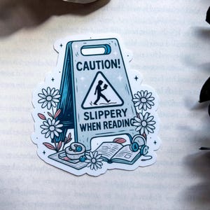 May include: A sticker featuring a blue and white caution sign with the text "CAUTION! SLIPPERY WHEN READING." The sign depicts a person walking with a book. Daisies, a book, headphones, and a cup of tea surround the sign.