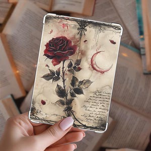 May include: A phone case featuring a dark red rose with green leaves and thorns. The design includes a crescent moon and handwritten script on a vintage-style background. The case is held in a hand.