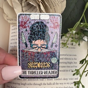 May include: A rectangular holographic sticker featuring an illustration of a person with a dark bun hairstyle and blue glasses reading a book. The book has red drips, two knives, and a yellow crime scene tape graphic with the text THE THRILLER READER.
