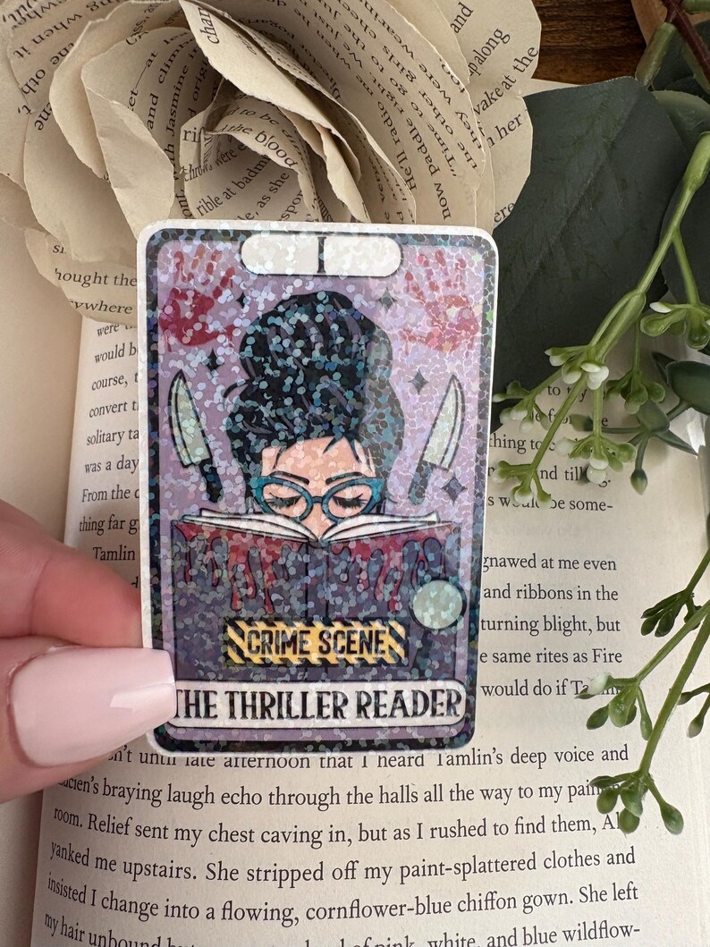 May include: A rectangular holographic sticker featuring an illustration of a person with a dark bun hairstyle and blue glasses reading a book. The book has red drips, two knives, and a yellow crime scene tape graphic with the text THE THRILLER READER.