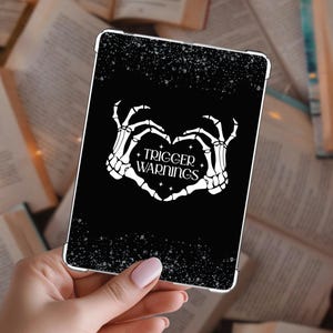May include: Black tablet case with a white skeleton hand heart design and the words "TRIGGER WARNINGS". The case has a speckled pattern and a silver border. The background is a collection of open books.