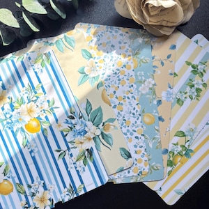 Può includere: Una collezione di fogli di carta fantasia a tema limoni e fiori. I disegni includono strisce blu e bianche, sfondi gialli e composizioni floreali con limoni. Le carte sono disposte a ventaglio, mostrando la varietà dei motivi.