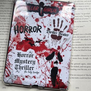 May include: A clear protective case for an e-reader, featuring a horror-themed design. The case is white with red blood splatter and various stickers with text like "HORROR" and "Thriller Reader".