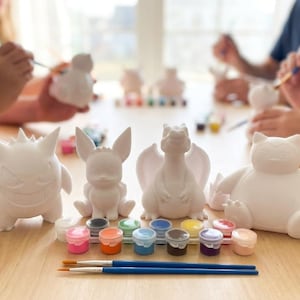 以下が含まれることがあります： ゲンガー、イーブイ、リザードン、カビゴンなど、白のセラミックフィギュアのコレクション。様々な色の絵の具と筆がフィギュアの前に配置されており、楽しく創造的な活動を示唆しています。背景には、同様のフィギュアをペイントしている人々がいます。