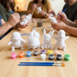 以下が含まれることがあります： 人気キャラクターの白く塗装されていないフィギュアのコレクション。塗装する準備ができています。明るい木製のテーブルに配置され、さまざまな色の小さなペイントポットと2本のペイントブラシが並んでいます。背景には、同様のフィギュアをペイントしている人々がいます。