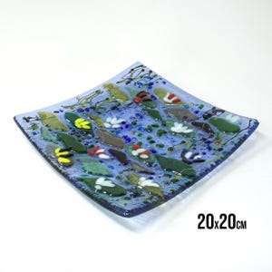 Könnte beinhalten: Eine quadratische Glasservierplatte mit blauem Hintergrund und einem Mosaikdesign aus grünen Blättern, weißen Blüten und bunten Akzenten. Die Platte misst 20x20 cm.