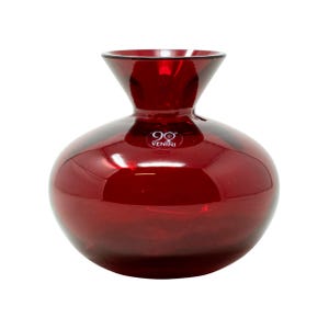 May include: A ruby red glass vase with a rounded body and a flared neck. The vase has a smooth, glossy finish and the text "VENINI" is visible on the front. This decorative vase is suitable for displaying flowers or as a standalone art piece.