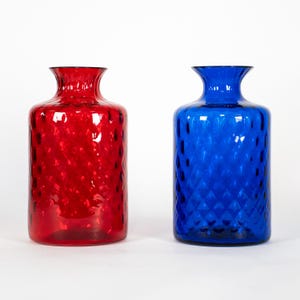 Könnte beinhalten: Zwei Glasvasen, eine rote und eine blaue, mit einem strukturierten Rautenmuster. Die Vasen haben eine zylindrische Form mit einem ausgestellten Hals. Die rote Vase befindet sich links und die blaue Vase rechts.