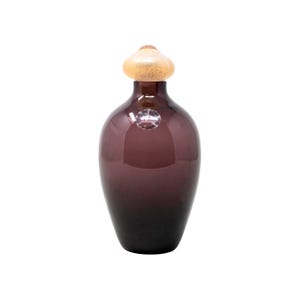 May include: A dark purple glass bottle with a bulbous body and a rounded top. The bottle has a gold-colored stopper. The glass has a glossy finish. A small, round sticker is affixed to the bottle.
