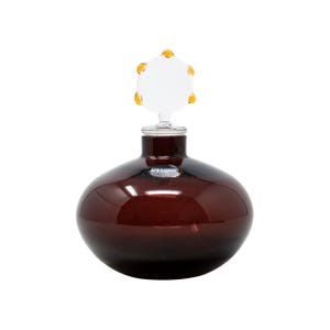 May include: A dark brown glass bottle with a round body and a clear stopper. The stopper has a white center with six yellow accents. The bottle is likely for perfume or other liquids.
