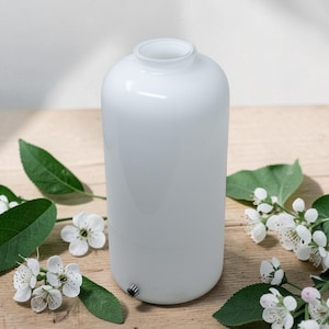 May include: A cylindrical white glass diffuser with a smooth, matte finish and a small circular metallic button near the base. The vessel features an open top and is surrounded by several sprigs of white blossoms with green leaves.