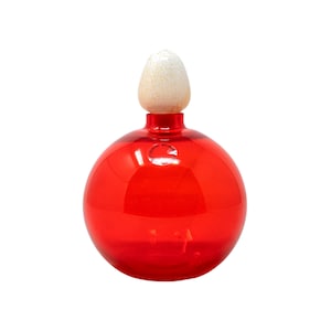 May include: A round, red glass bottle with a white and tan stopper. The bottle has a glossy finish and a small, circular label. The stopper is cone-shaped and textured. The bottle is likely for decorative purposes.