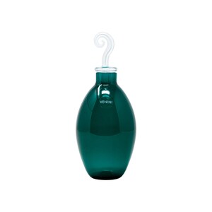 May include: A teal-colored glass bottle with a clear, swirled handle. The bottle has a rounded shape and a narrow neck. The word "VENINI" is printed on the bottle. The bottle is likely used for decorative purposes.