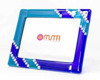 May include: A blue and turquoise glass picture frame with white decorative accents. The frame has a beveled edge and a clear acrylic stand. The frame is decorated with small white flowers. The frame is made by "Muma, Idee e Colori da Venezia".