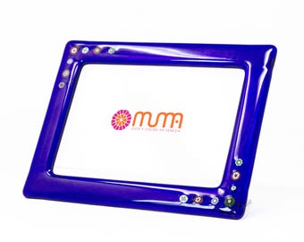 May include: A blue glass picture frame with colorful glass accents. The frame has a beveled edge and a smooth finish. The frame is decorated with small, colorful glass beads. The frame is made in Venice, Italy. The text "MUMA, GIOIE E COLORI DA VENEZIA" is visible in the frame.