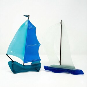 May include: Two glass sailboat sculptures. One has a blue and turquoise sail, the other has a clear sail. Both have dark blue hulls and masts. The sculptures are on a white background.
