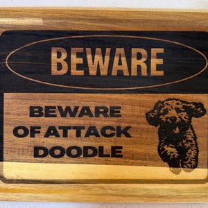 May include: A rectangular wooden tray with a natural grain finish featuring a dark engraved design. The text reads BEWARE at the top and BEWARE OF ATTACK DOODLE below, accompanied by a detailed laser-engraved illustration of a curly-haired dog.