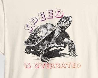 Op de afbeelding: Een crèmekleurig T-shirt met een zwart-witte illustratie van een schildpad en de tekst "SPEED IS OVERRATED" in roze en rood.