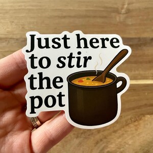 May include: A sticker with the text "Just here to stir the pot" in a bold, black font. The sticker features a cartoon illustration of a black pot filled with soup, with a wooden spoon. The sticker is on a wooden surface.