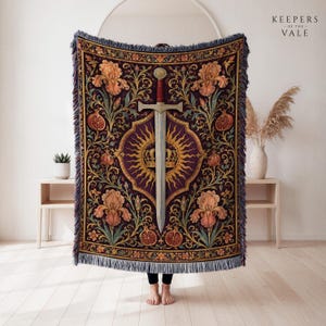 May include: A woven throw blanket featuring a central silver sword and a golden crown set against a sunburst motif. The deep purple background is decorated with orange iris flowers and pomegranates. The edges are finished with blue fringe.