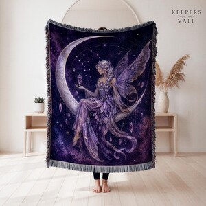 May include: A woven throw blanket featuring a purple and dark blue cosmic background with a fairy sitting on a crescent moon. The fairy has wings and holds a crystal. The design includes hanging crystals and stars. The text KEEPERS OF THE VALE is visible.