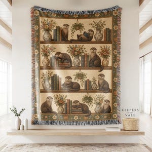Könnte beinhalten: Eine gewebte Wohndecke mit einem wiederkehrenden Regaldesign, das Illustrationen von Ottern, Bücherstapeln und Blumenvasen zeigt. Der Rand hat ein Blumenmuster und Fransen. Der Text "KEEPERS OF THE VALE" ist unten rechts aufgedruckt.