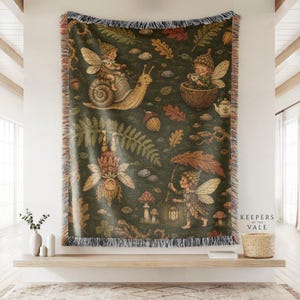 May include: A woven throw blanket with a dark green background featuring a repeating pattern of winged fairies, snails, acorns, mushrooms, and autumn leaves. The blanket has fringed edges and the text Keepers of the Vale printed in the corner.