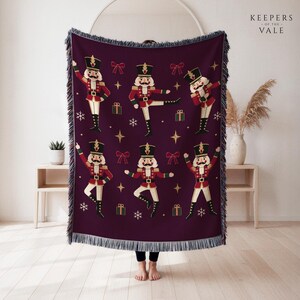 May include: A deep purple woven throw blanket featuring a repeating pattern of nutcracker figures in red and gold uniforms, interspersed with small red bows, green gift boxes, and white snowflakes, finished with a fringed edge on all sides.