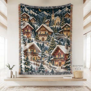 Könnte beinhalten: Eine gewebte Wohndecke mit einer winterlichen Dorfszene, verschneiten Häusern, Tannenbäumen und einer Kirche unter einem Sternenhimmel. Die Decke hat Fransen an den Rändern und den Schriftzug Keepers of the Vale in der unteren rechten Ecke.