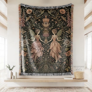 May include: A woven tapestry blanket featuring a dark background with two winged fairy figures holding hands. The design includes intricate botanical elements, mushrooms, flowers, and a central eye motif. The text "KEEPERS OF THE VALE" is printed in the corner.