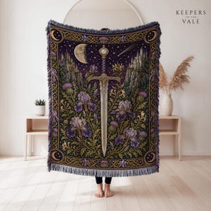 May include: A woven throw blanket featuring a central sword surrounded by purple irises and thistles against a dark purple night sky with a crescent moon, stars, and castles on cliffs. The border has a gold Celtic knot pattern with fringed edges.