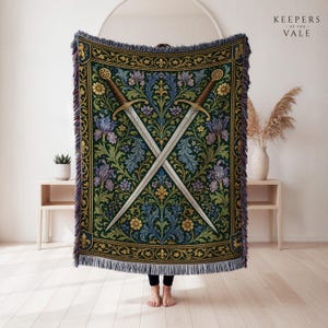 May include: A woven throw blanket featuring a dark green background with a symmetrical pattern of crossed swords, purple irises, and gold floral motifs. The rectangular textile has a decorative border and fringed edges on the top and bottom sides.