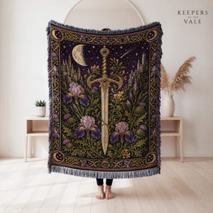 May include: A woven throw blanket featuring a central sword surrounded by purple iris flowers, green thistles, and a crescent moon against a dark starry sky. The rectangular blanket has a fringed edge and a decorative border with gold accents.