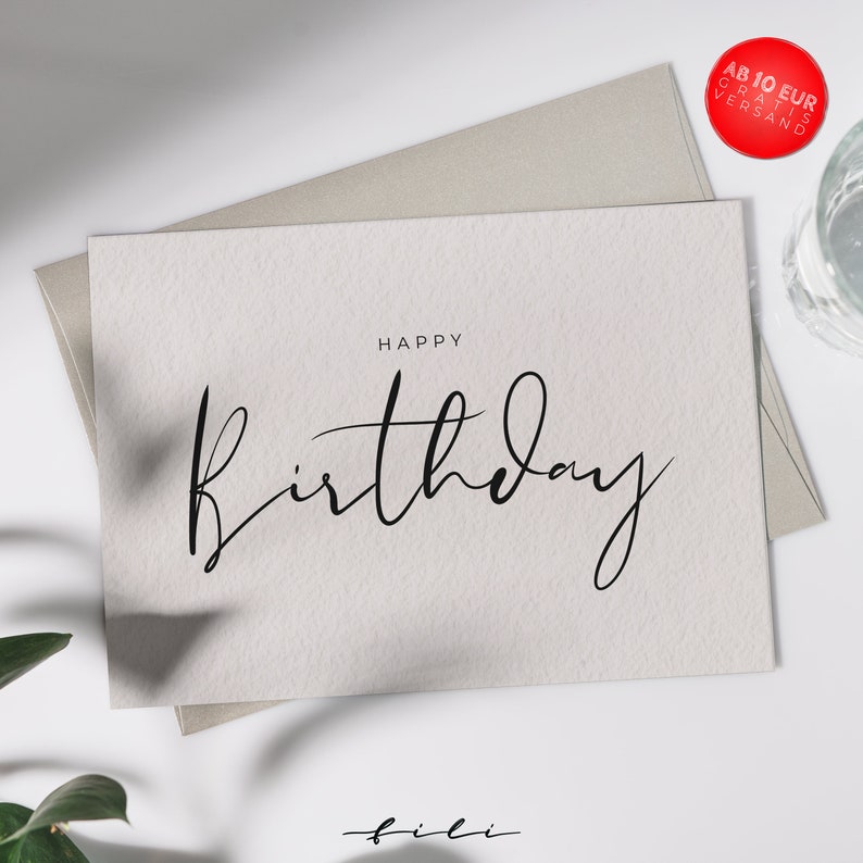 May include: A white card with the text "Happy Birthday" written in black cursive script. The card is on a light gray surface with a green plant in the bottom left corner.