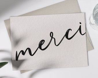May include: A white card with the word "merci" written in black cursive script. The card is on a light gray surface.